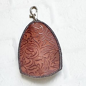 May include: A teardrop-shaped keychain featuring a brown leather surface with an embossed floral pattern. The edges are finished with dark stitching, and a silver-toned metal lobster clasp is attached to the top for secure fastening.