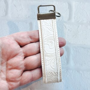 May include: Cream-colored key fob with a silver-tone metal key ring and clasp. The fob features an embossed floral pattern and is bordered by a stitched edge. The key fob is designed to hold keys and is a stylish accessory.