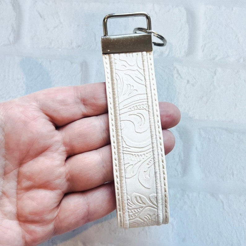 May include: Cream-colored key fob with a silver-tone metal key ring and clasp. The fob features an embossed floral pattern and is bordered by a stitched edge. The key fob is designed to hold keys and is a stylish accessory.