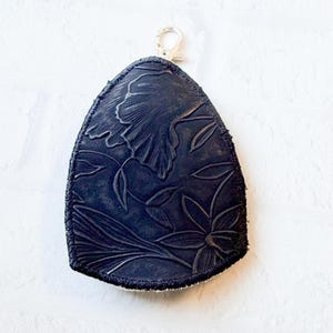 May include: A dark blue leather key fob with a floral design. The fob has a silver-colored clasp at the top for attaching to a keychain. The floral pattern is embossed into the leather.