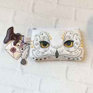 May include: A white zipped pouch featuring an embroidered owl face with yellow eyes and intricate grey scrollwork. Attached to the zip pull are three embroidered charms: a brown hat, a white envelope with a red seal, and a white quill with ink.