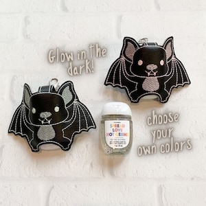 May include: Two black bat-shaped holders with embroidered details and silver metal clips, alongside a clear plastic bottle of hand sanitizer labeled 'Spread Love Not Germs' and 'Sea Salt & Citrus 1 fl oz / 29 ml'. Text reads 'Glow in the dark!' and 'choose your own colors'.
