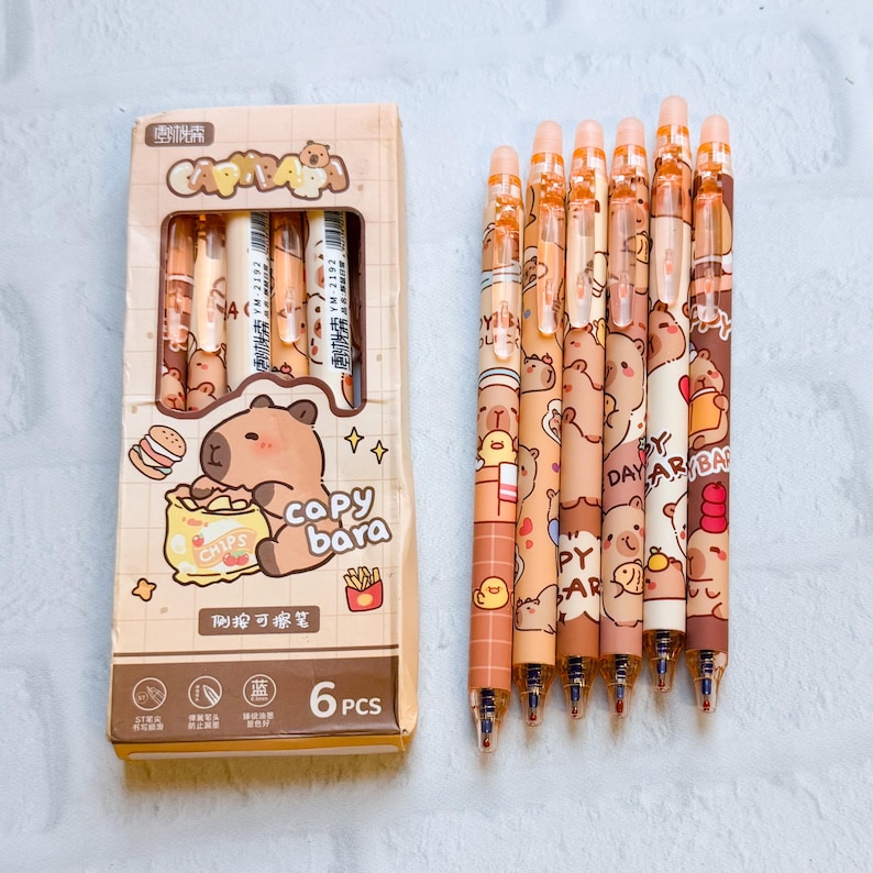 May include: A set of six retractable ballpoint pens with a cute bear design in a box. The pens are various shades of brown and beige, with the box featuring a bear eating chips. The box reads "CAPYBARA" and "6 PCS".
