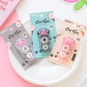 May include: Three pink and white cat paw-shaped tape dispensers with the text "Cat Paws" and "TG-81694A" on each. The dispensers are on a white surface.