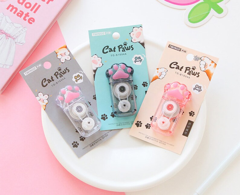 May include: Three pink and white cat paw-shaped tape dispensers with the text "Cat Paws" and "TG-81694A" on each. The dispensers are on a white surface.