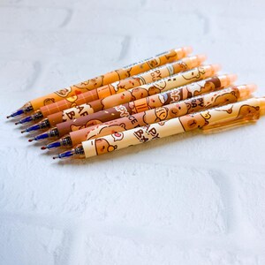 May include: Seven gel pens with brown and tan bear designs. The pens have blue ink tips and are arranged diagonally on a white brick background. The pens have a variety of bear illustrations and the text "HAPPY".