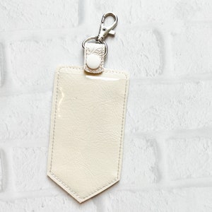 May include: A cream-coloured rectangular badge holder with a pointed bottom edge, featuring a clear plastic front window, white stitching, a matching cream-coloured tab with a white snap button, and a silver-toned metal swivel lobster claw clasp.