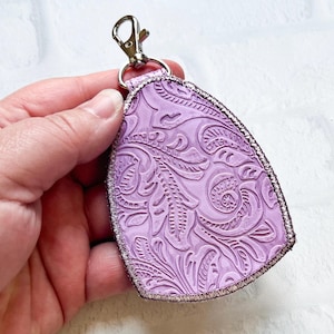 May include: A lavender-colored key fob with a silver-colored clasp. The key fob has a detailed embossed floral pattern and a silver-colored stitched border. The key fob is held in a hand.