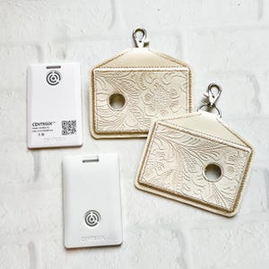May include: An overhead shot shows two cream-colored keychains with floral embossed designs and silver-tone clasps, alongside two white rectangular Centegix devices. One device has the Centegix logo, model information, and a QR code.
