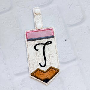 May include: A pencil-shaped keyring tag featuring a white embossed floral pattern, a pink eraser section, a black embroidered letter T, and a brown cork-textured pencil tip. The tag includes a white snap fastener on a vertical tab.