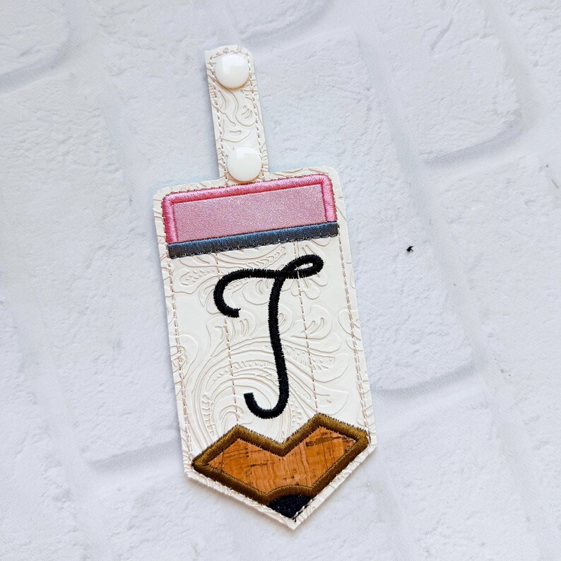 May include: A pencil-shaped keyring tag featuring a white embossed floral pattern, a pink eraser section, a black embroidered letter T, and a brown cork-textured pencil tip. The tag includes a white snap fastener on a vertical tab.