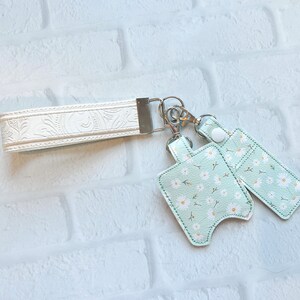 May include: A white wristlet keychain with a floral embossed design, connected to two mint green hand sanitizer holders. The holders feature a daisy pattern and silver hardware.