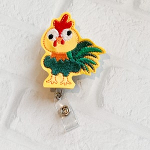 May include: A badge reel featuring a yellow embroidered chicken with a red comb, orange neck feathers, and dark green wings. The chicken has large white eyes with black pupils and a small orange beak. A clear plastic strap with a metal snap is attached.