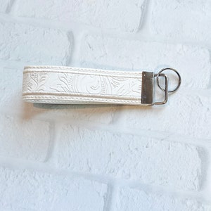 May include: A white wristlet keychain with a floral embossed design. The keychain features a silver-colored metal clasp and key ring. The wristlet is made of a textured material and is against a white brick background.