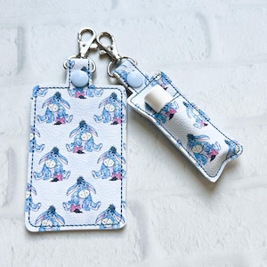 May include: Two white accessories with a cartoon donkey pattern. One is a card holder with a snap closure and a silver clip. The other is a lip balm holder with a silver clip and a lip balm inside.