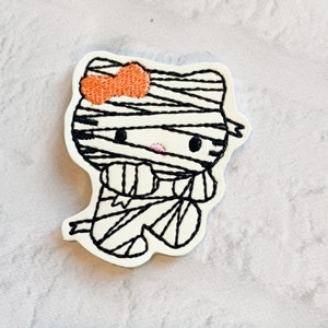 May include: A white embroidered patch of a cartoon mummy cat with a pink nose and orange bow. The cat is wearing a white bandage with black stitching.