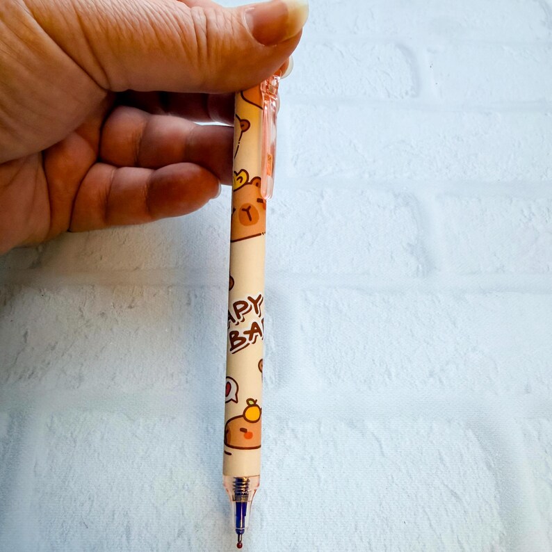 May include: A beige pen with a repeating pattern of brown bears and the text "HAPPY BABY". The pen has a gold-colored tip and a blue ink cartridge. The pen is held by a hand.