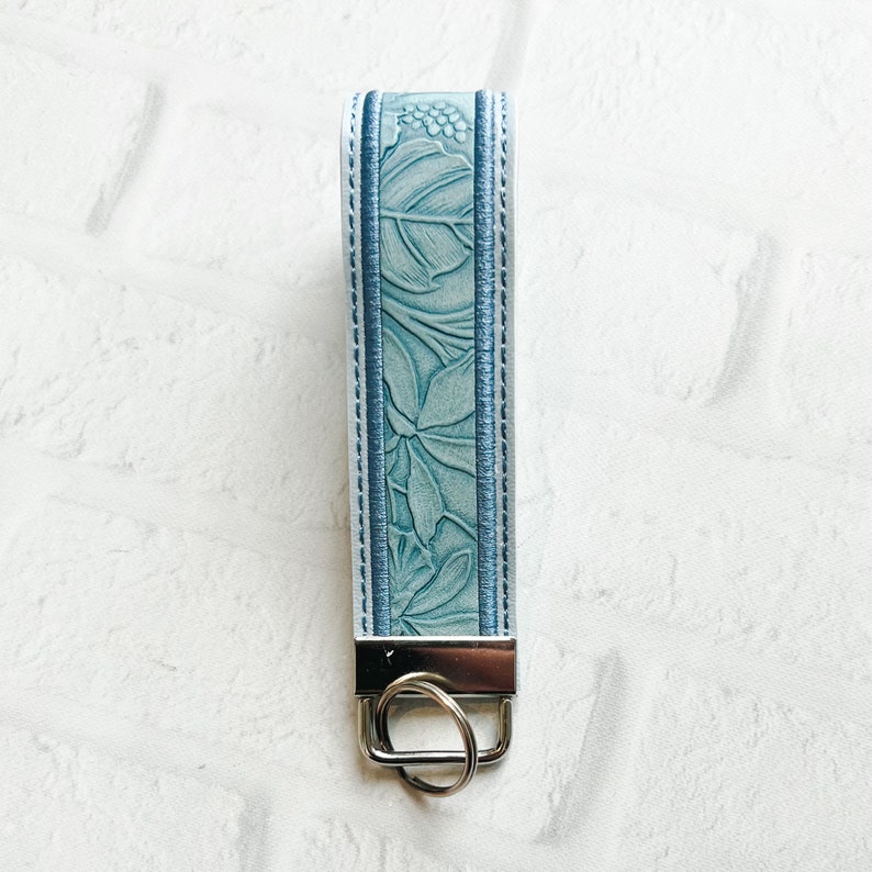 May include: A light blue keychain strap featuring an embossed floral pattern with darker blue stitched borders. The strap is attached to a silver-toned metal hardware clamp with a circular split ring at the bottom for holding keys securely.
