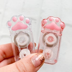 May include: Two pink and clear correction tape dispensers shaped like a cat paw print. The dispensers have glitter embedded in the plastic. The text "Correction Tape" is printed on the side of each dispenser.