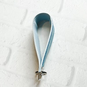 May include: A light blue wristlet keychain featuring a smooth interior and a patterned exterior with white stitching along the edges. The strap is looped and secured at the base with a silver-toned metal clasp and a circular split ring attachment.