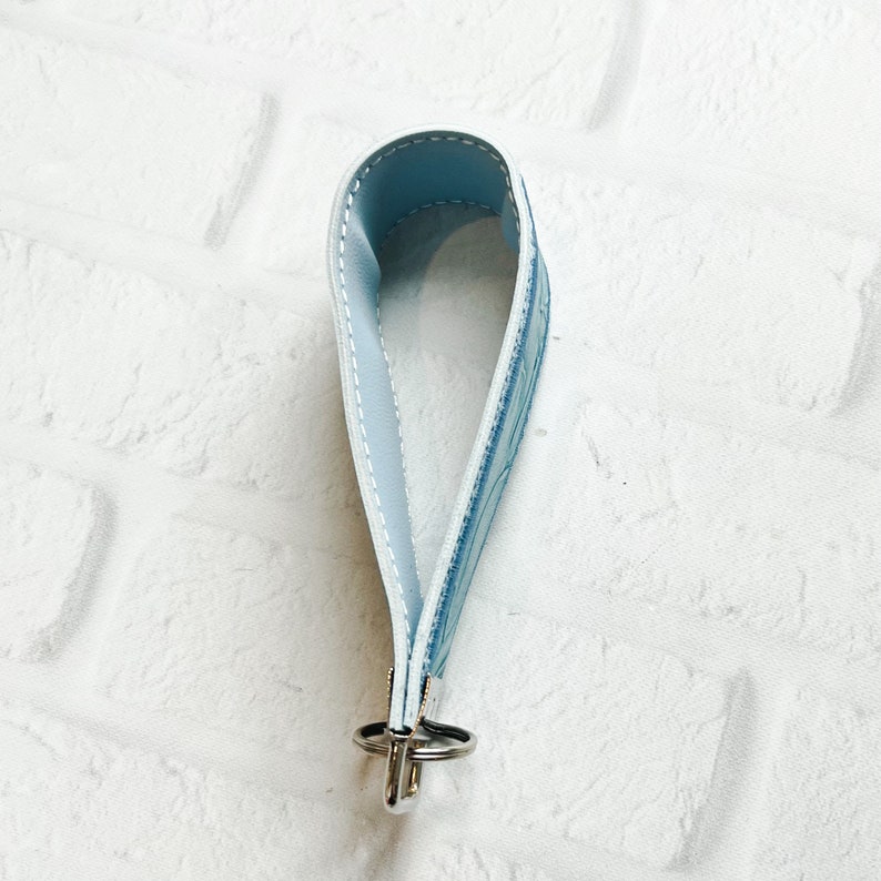 May include: A light blue wristlet keychain featuring a smooth interior and a patterned exterior with white stitching along the edges. The strap is looped and secured at the base with a silver-toned metal clasp and a circular split ring attachment.