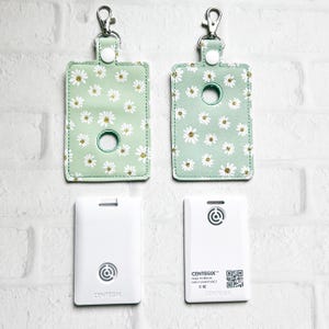 May include: Two white keychains with a green daisy print design. The keychains have a small hole for attaching a dog waste bag dispenser. The keychains are attached to white cards with the text "CENTEGIX" and a logo.