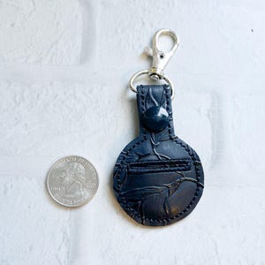 May include: A black leather keychain with a silver clasp and a decorative embossed design. The keychain has a button closure and a small pocket. A U.S. quarter is next to the keychain for size comparison.
