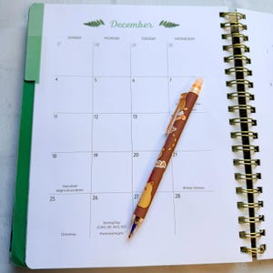 May include: A spiral-bound calendar open to December, with a brown pen resting on the pages. The pen has a gold clip and a brown barrel with a cartoon design. The calendar has a green spine and gold spiral binding.