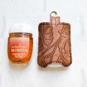 May include: A clear bottle of Sunshine Mimosa anti-bacterial hand gel next to a brown leather hand sanitizer holder. The holder features a floral design and a silver-colored clasp. The hand gel bottle is 1 fl oz / 29 ml.