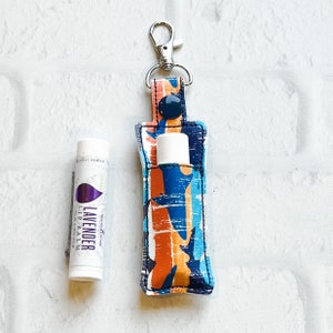 May include: A lip balm holder with a blue, orange, and white abstract pattern, attached to a silver clip. Next to it is a lavender lip balm tube. The holder has a navy blue snap closure and holds the lip balm securely.
