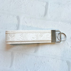 May include: Ivory-colored wristlet keychain with a floral embossed design. The keychain features a silver-tone metal clasp and key ring. The wristlet is made of a durable material and is designed to be worn around the wrist for easy access to keys.
