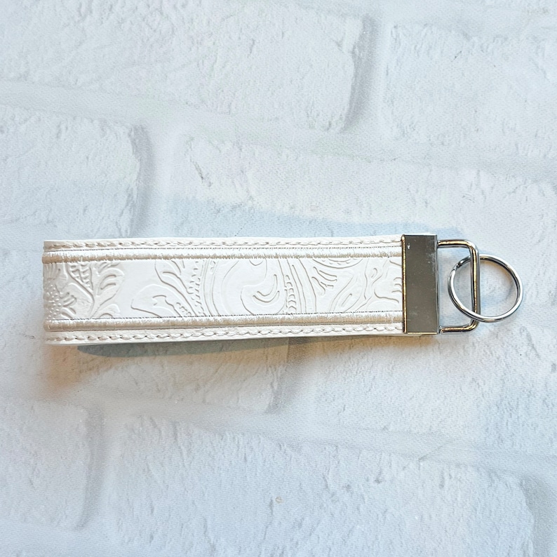 May include: Ivory-colored wristlet keychain with a floral embossed design. The keychain features a silver-tone metal clasp and key ring. The wristlet is made of a durable material and is designed to be worn around the wrist for easy access to keys.