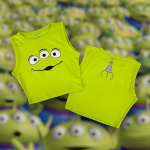 Puede incluir: Un top corto sin mangas de color verde lima con una cara de alienígena de tres ojos en la parte delantera y una garra de una máquina de garras en la parte trasera. El texto "RUYA" está impreso en la parte delantera de la camiseta.