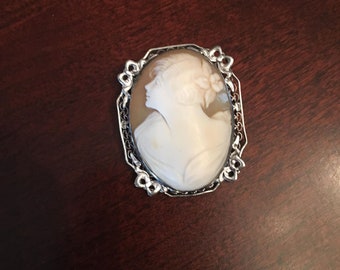May include: A silver cameo brooch with a white cameo of a woman's profile. The cameo is set in a silver frame with a decorative design.