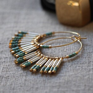 May include: Gold hoop earrings with turquoise and silver beads hanging from the bottom. The earrings are on a light brown fabric background.
