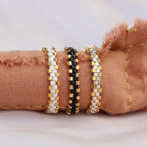 May include: Three beaded rings in black, white, and silver with gold accents. The rings are stacked on a peach-colored fabric.