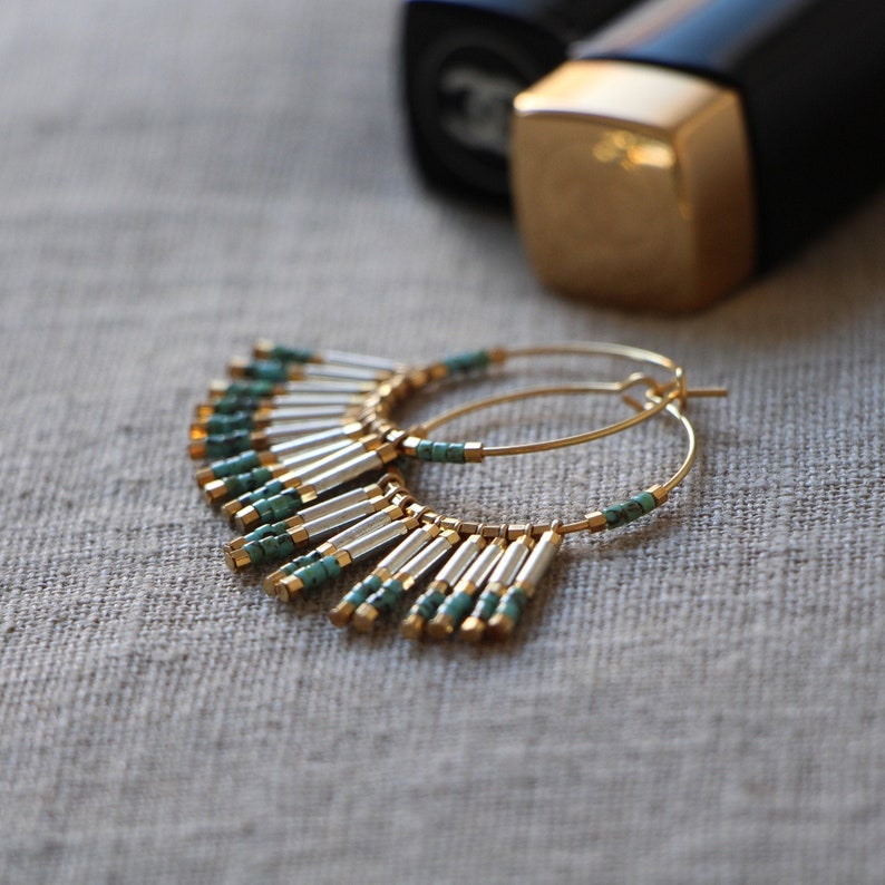 May include: Gold hoop earrings with turquoise and silver beads hanging from the bottom. The earrings are on a light brown fabric background.