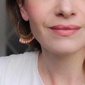 May include: A pair of gold hoop earrings with pink and green beaded fringe. The earrings are worn by a person with light skin and blonde hair.