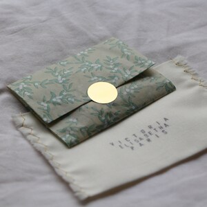 May include: A small, white gift bag with a green floral pattern and a gold circle sticker. The bag is folded over and sitting on a white fabric surface. The text "VICTORIA ELISABETH PARIS" is printed on the fabric surface.