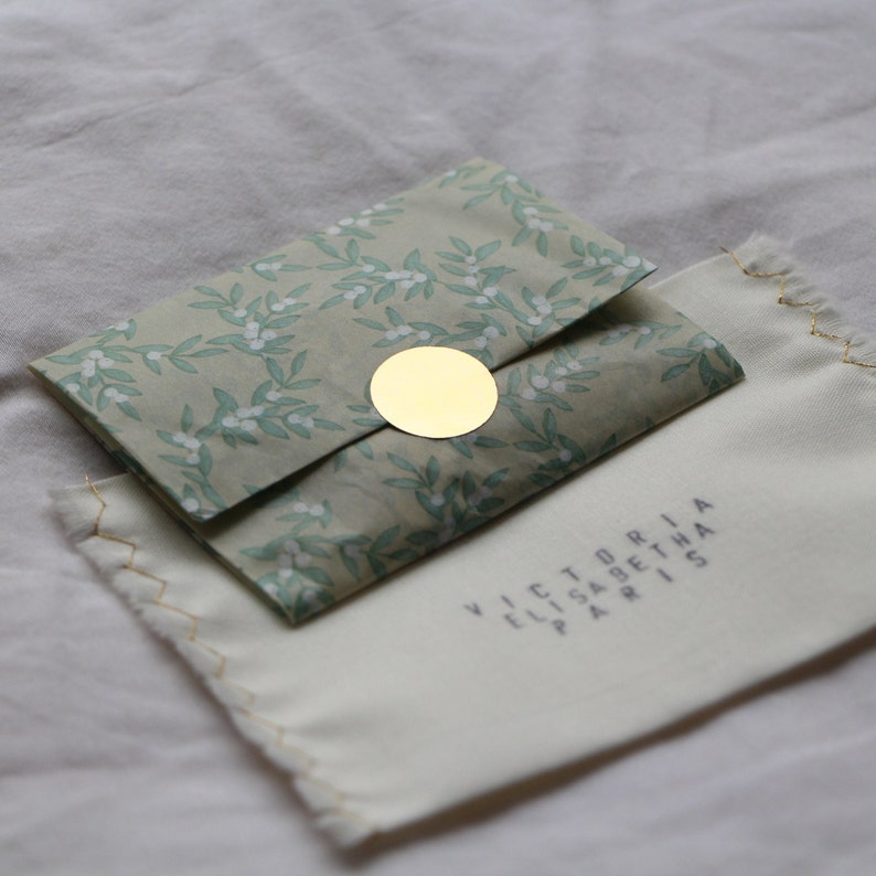 May include: A small, white gift bag with a green floral pattern and a gold circle sticker. The bag is folded over and sitting on a white fabric surface. The text "VICTORIA ELISABETH PARIS" is printed on the fabric surface.