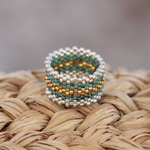 May include: A turquoise, silver, and gold beaded ring. The ring is made with small square beads and has a geometric pattern.