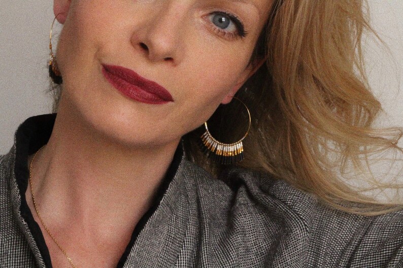 May include: A woman wearing a grey blazer with gold hoop earrings featuring black and white beads dangling from them. The earrings are a statement piece and add a touch of elegance to the outfit.