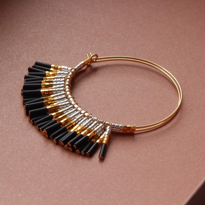 May include: Gold hoop earrings with a black and silver beaded fringe. The fringe is made of small, cylindrical beads that hang down from the hoop.