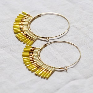 May include: A pair of gold hoop earrings with yellow and white beaded fringe. The fringe is made of small, cylindrical beads that are arranged in a graduated pattern.