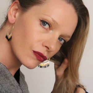 May include: A woman with blonde hair is wearing a pair of gold hoop earrings with black and white fringe. The earrings are dangling from her ears.