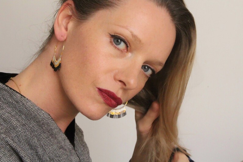 May include: A woman with blonde hair is wearing a pair of gold hoop earrings with black and white fringe. The earrings are dangling from her ears.