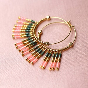 May include: Gold hoop earrings with pink and green beaded fringe. The fringe is made of small, round beads that hang from the hoop.