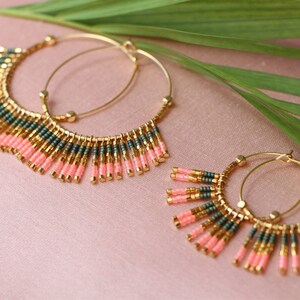 May include: A pair of gold hoop earrings with pink and green beaded fringe. The earrings are on a pink background with a green leaf.