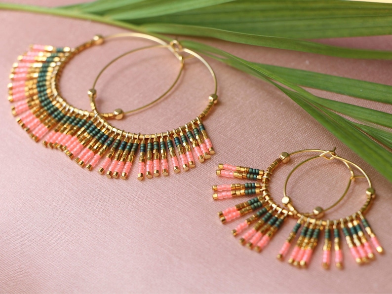 May include: A pair of gold hoop earrings with pink and green beaded fringe. The earrings are on a pink background with a green leaf.