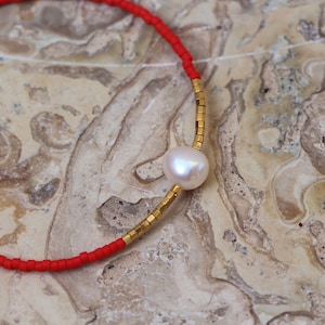 Op de afbeelding: Een rode en gouden kralen armband met een enkele witte parel bedel.